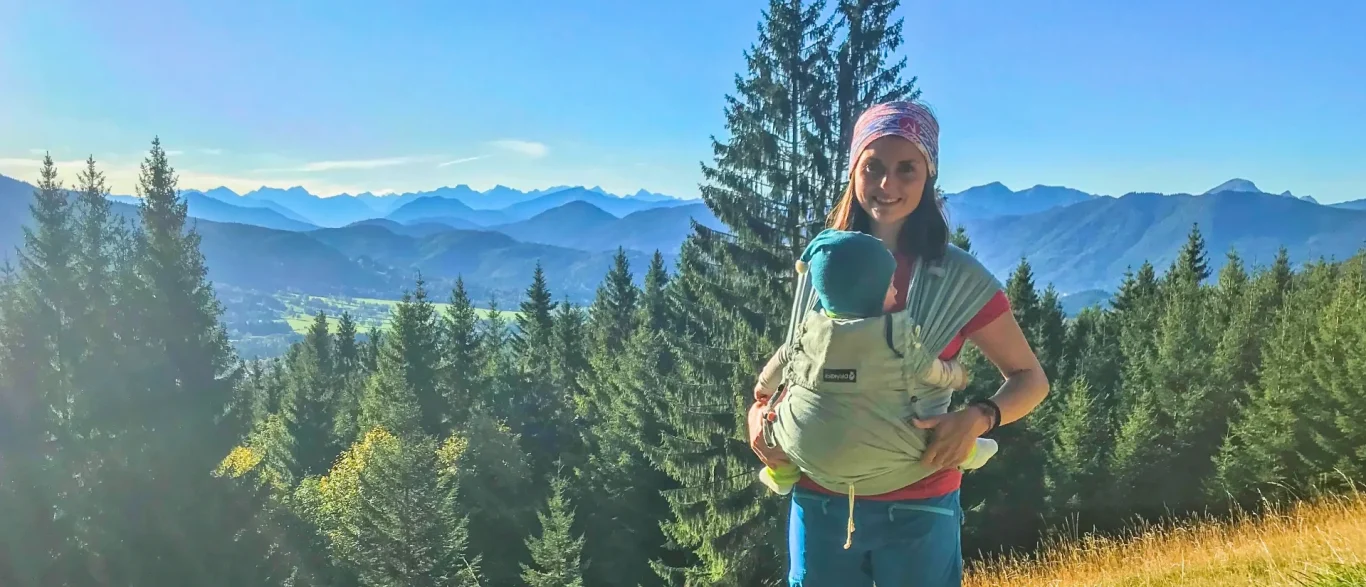 Titelbild Frau mit Baby-Trage vor einer bergigen Landschaft.