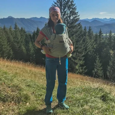 Hallo, ich bin Angelique... Angelique Rest mit Baby im Tragetuch, auf grüner Wiese vor einer Bergkulisse.
